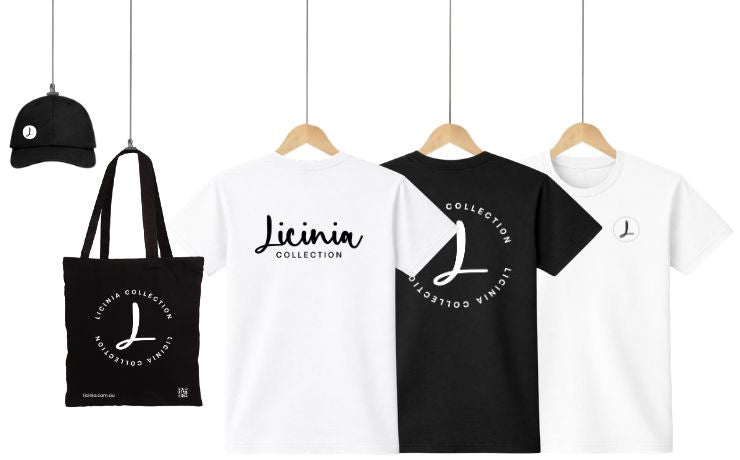 Flat lay display of black performance cap, black organic cotton tote bag, and black and white heavy t-shirts with custom Licinia logo and graphic motif prints, shown front and back on hangers against a clean white background, representing professional customer branding and custom apparel printing services.