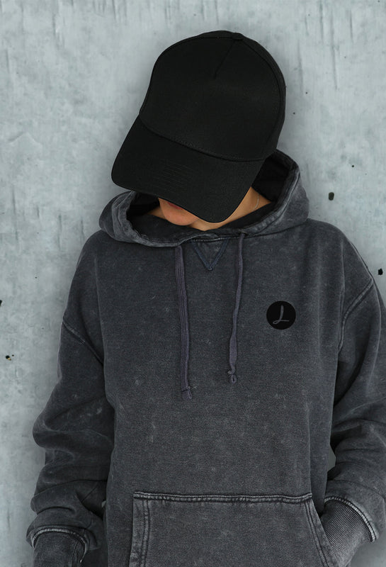 Female model wearing black vintage stone wash custom printed hoodie with Licinia logo and matching black A-frame cap, photographed against concrete wall for mobile hero slider showcasing branded fashion apparel.