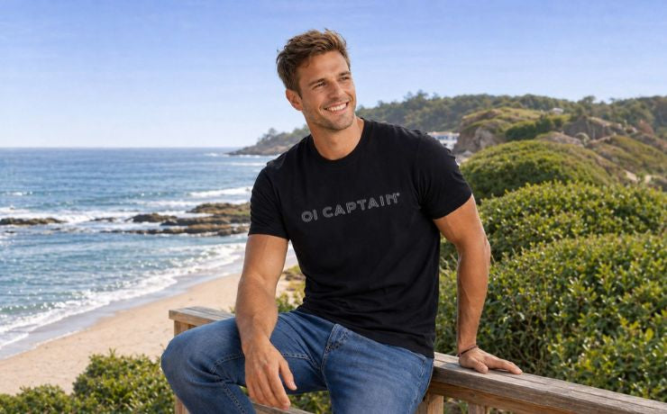 Male model in Oi Captain Eclipse Soft Style T-Shirt in black, half body front view sitting on a wooden fence at the beach with one knee raised smiling in a relaxed pose