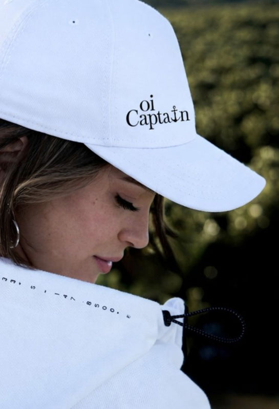 Close-up side profile of female model wearing Oi Captain Seascape Hoodie Jacket and Endless Summer white cap, highlighting Oi Captain logo on cap and Metung coordinates hoodie detail with natural green background featured1